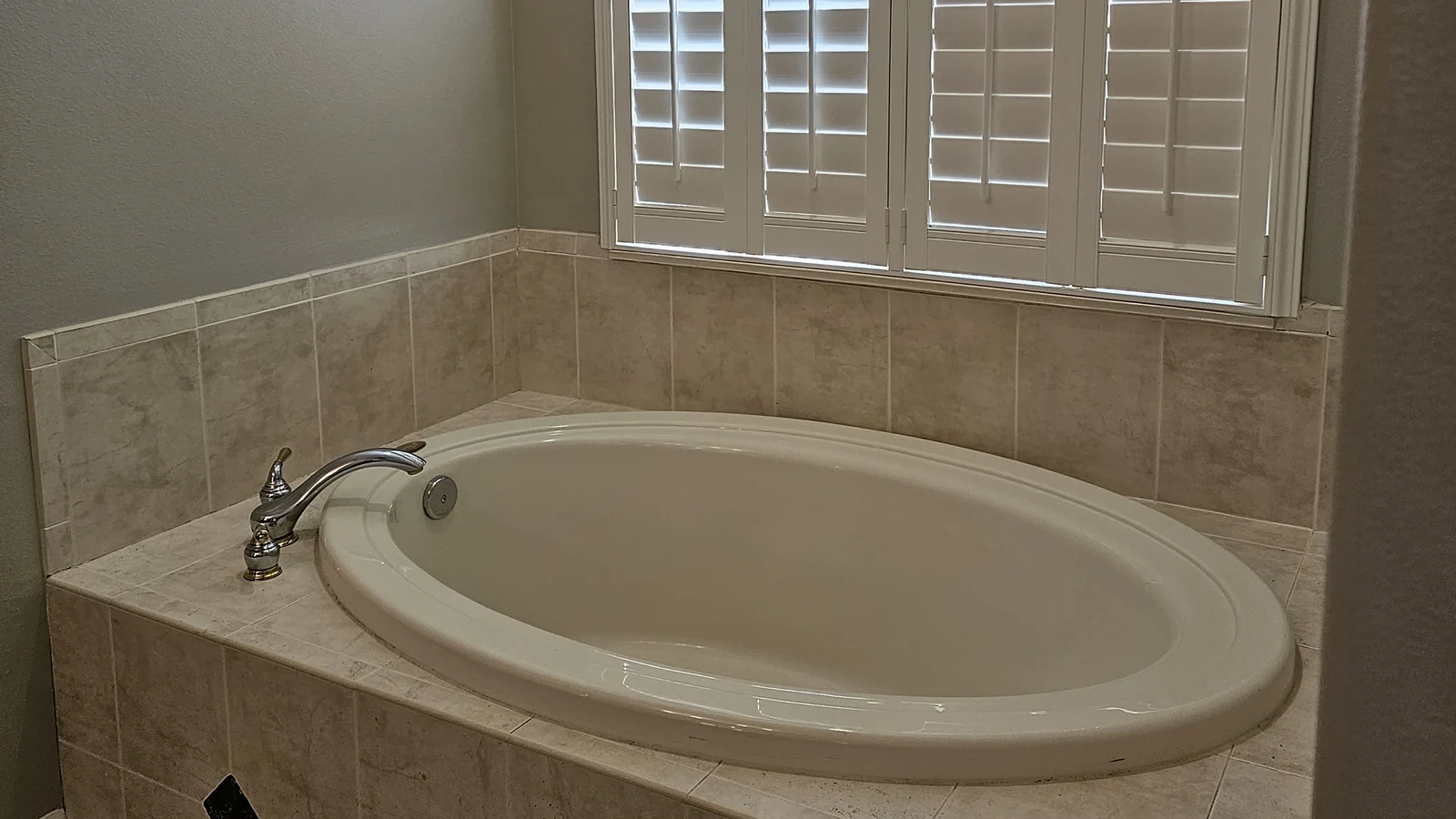 The Winter Park primary bath before remodeling: a built-in oval garden tub on a beige tile deck below a window with plantation shutters