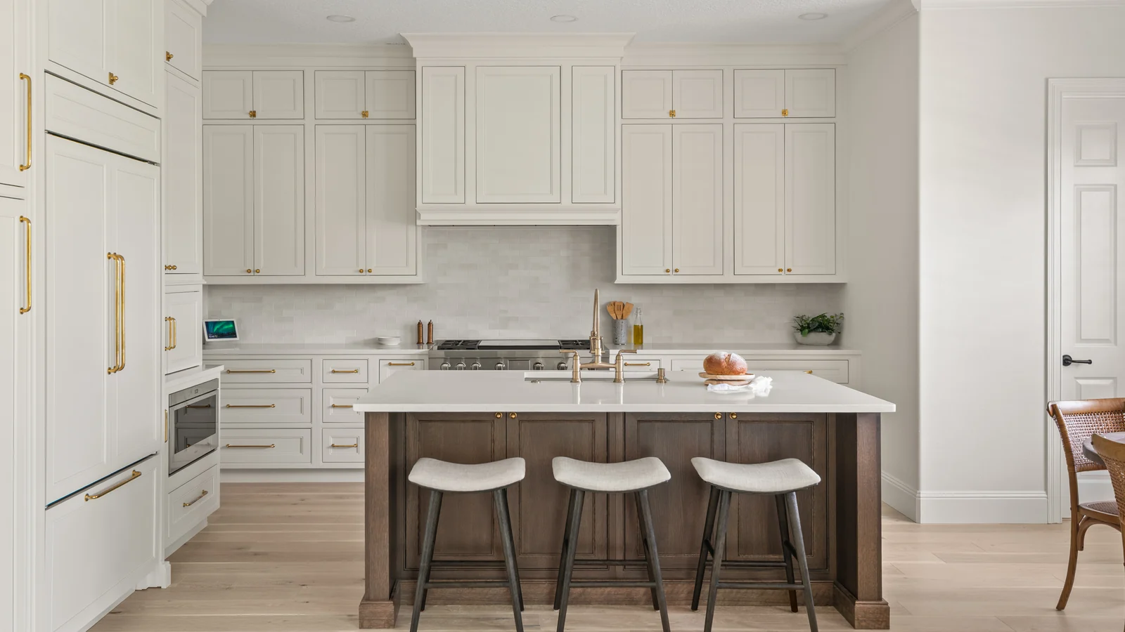 The same Winter Park kitchen after the whole-home remodel: ivory perimeter cabinets with brass hardware, paneled appliance fronts, and a stained-wood island under a white quartz top with three upholstered stools