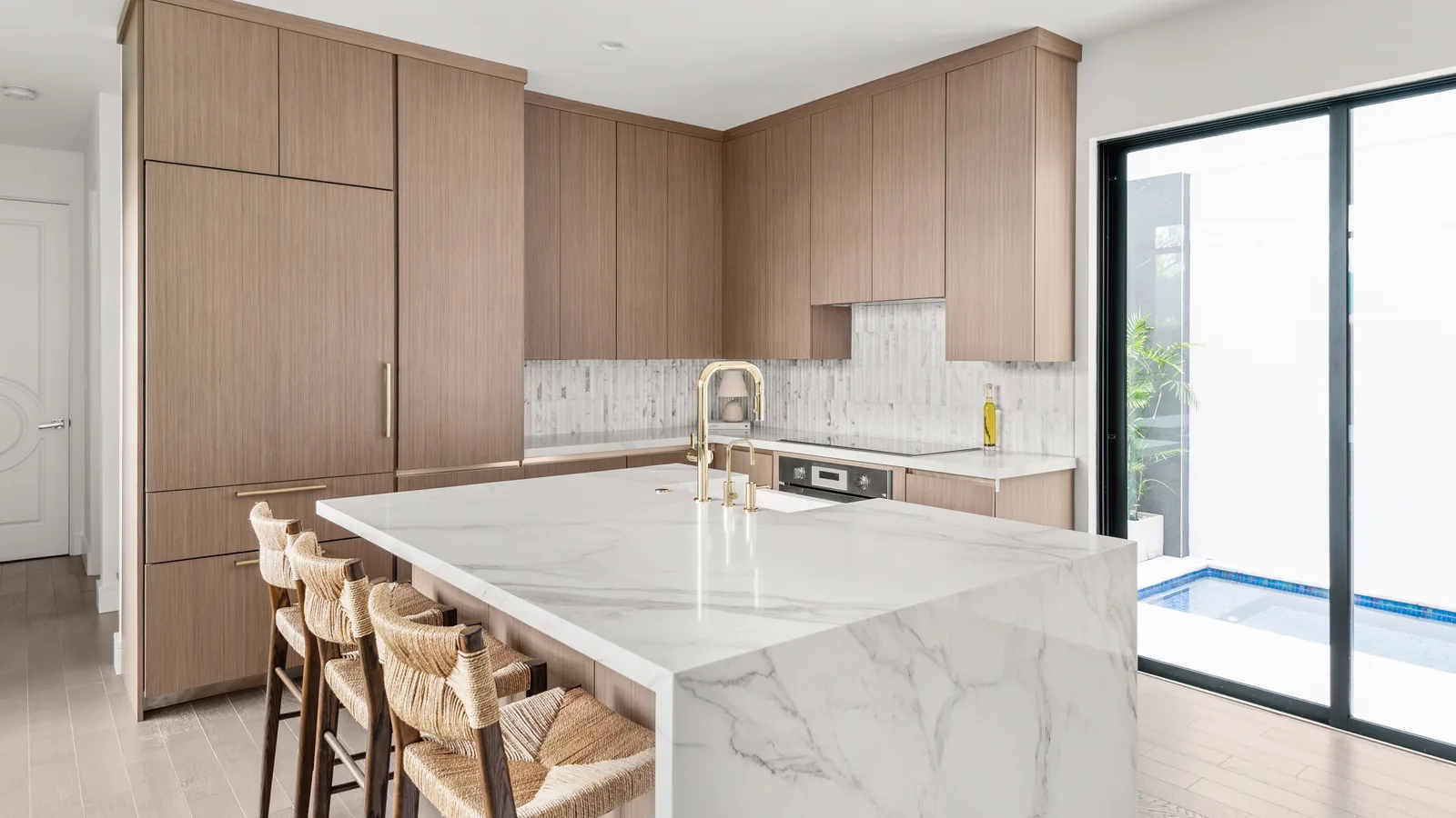 The built Winter Park kitchen photographed at completion: the same tall cabinet bank and marble-veined quartz waterfall island as the model, now in warm wood-grain slab fronts with woven stools, a brass faucet, and the pool just outside the sliding door