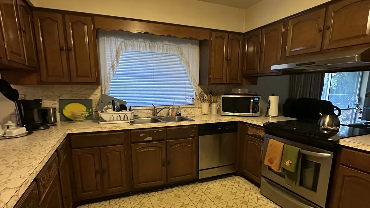 The same kitchen before remodeling: dark 1960s cabinets wrapping a closed galley under a small curtained window