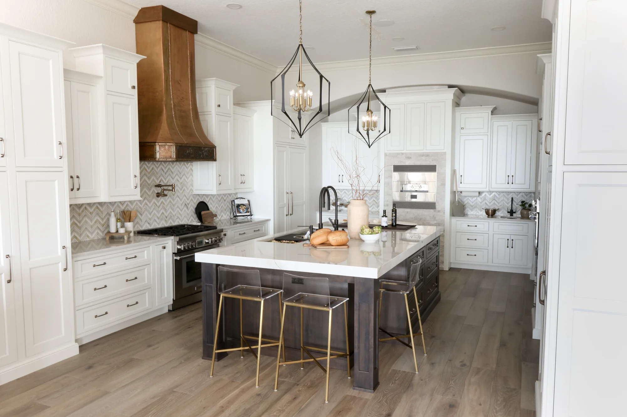 Windermere estate kitchen remodel &mdash; dark square island with brass-and-acrylic stools, chevron marble backsplash, and a commissioned hammered-bronze range hood