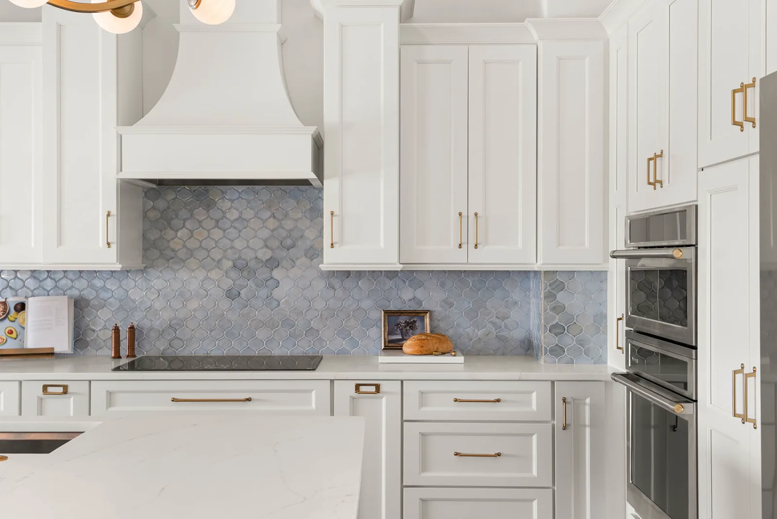 The blue arabesque tile backsplash running the full cooking wall, framed by white cabinetry with brass cup and bar pulls, the stacked stainless wall oven and microwave at right
