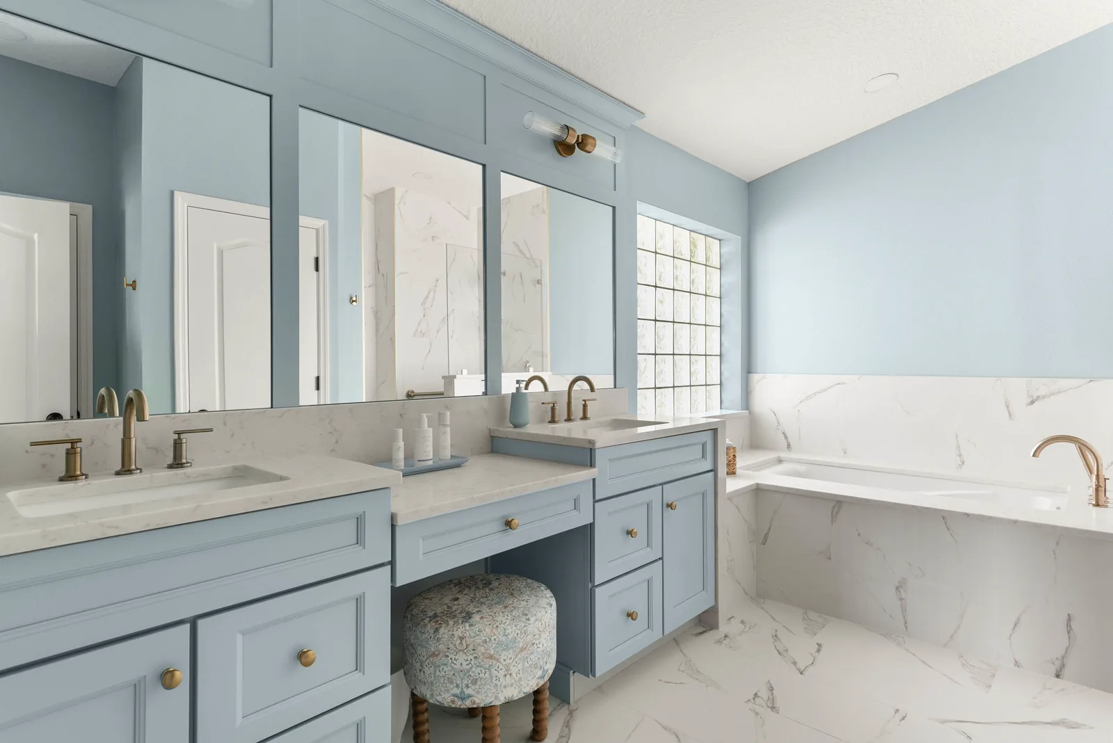 The remodeled Clermont primary bathroom: soft blue vanities with marble-veined counters flanking a seated makeup vanity, mirrors framed in blue millwork, and the marble-look tub deck at right