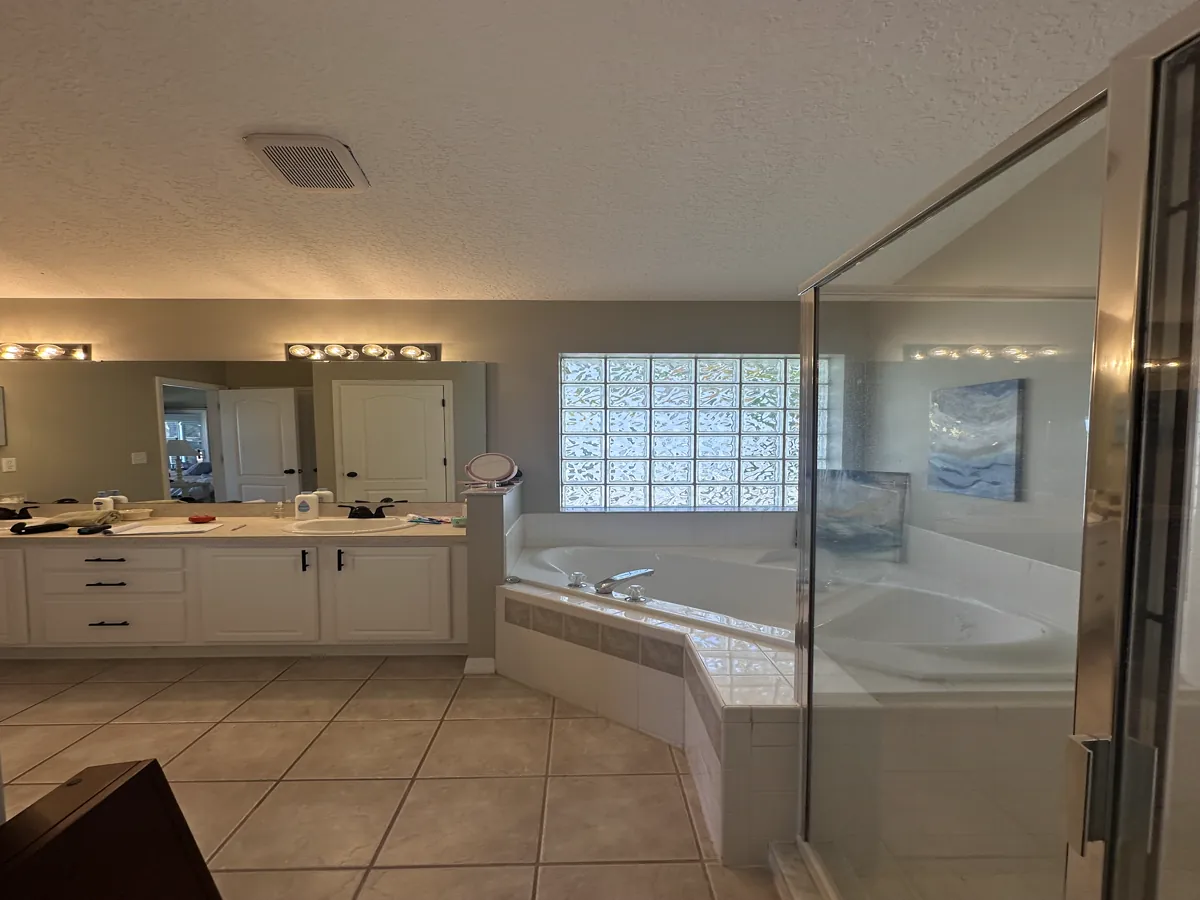 The primary bathroom before remodeling: a long white raised-panel vanity with a beige cultured-marble top and two drop-in oval sinks, a bulky corner drop-in tub in beige tile, and a framed chrome shower enclosure