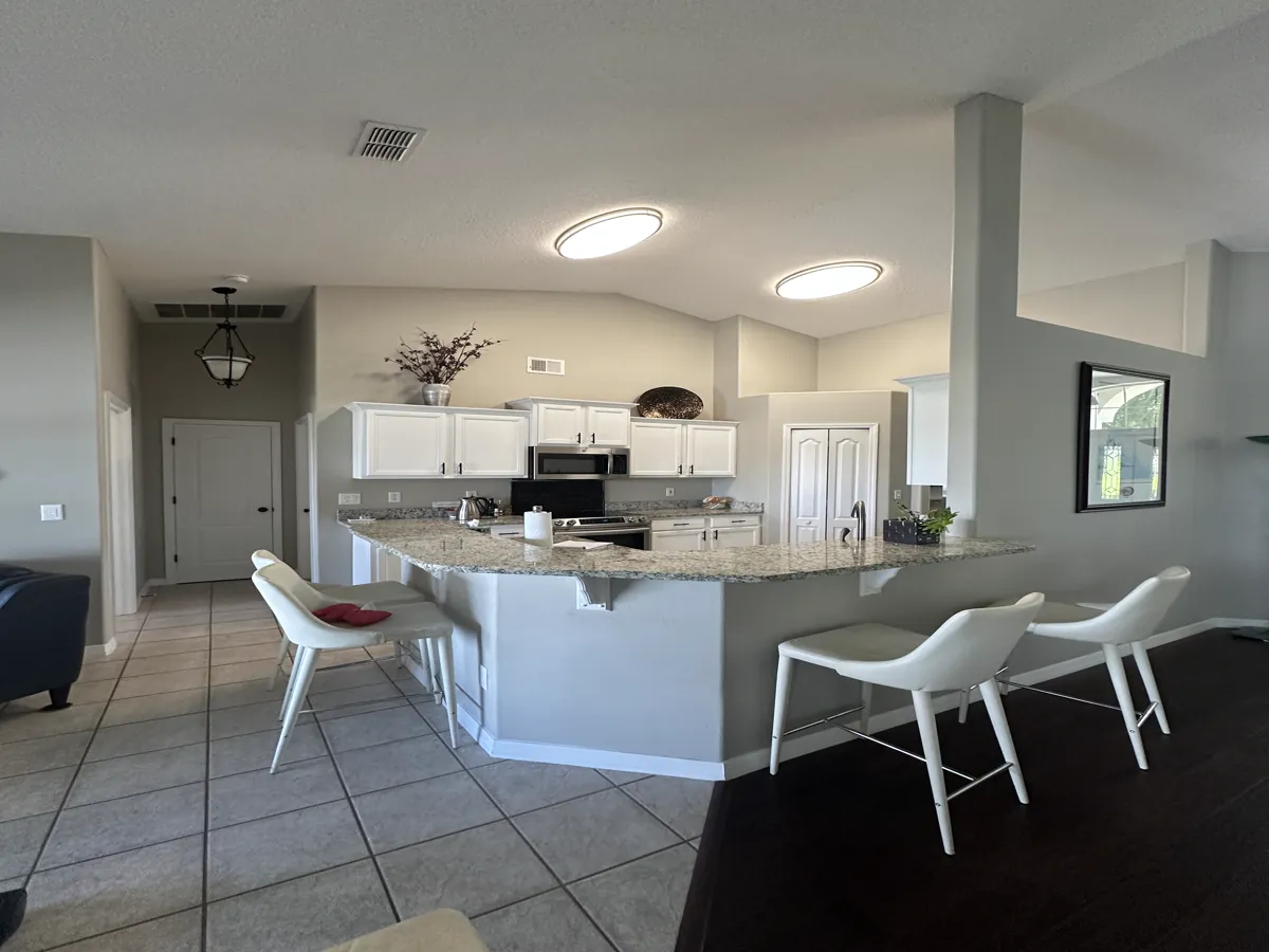 The Clermont kitchen before remodeling: an angled granite peninsula bar closing the kitchen off from the family room, white raised-panel cabinets, an over-range microwave, and beige ceramic tile floor