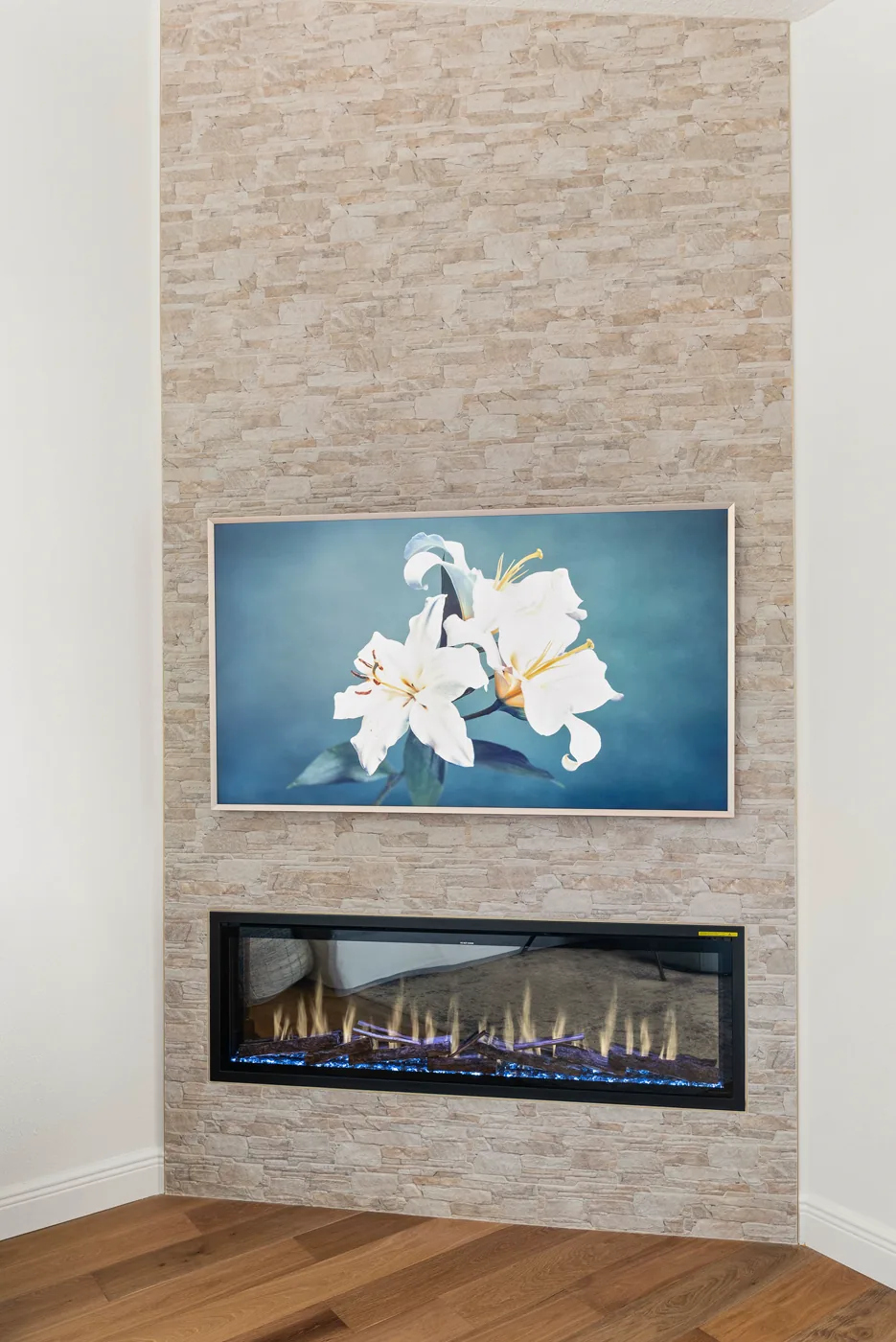 The stacked stone fireplace wall floor to ceiling, a linear firebox lit below a framed art panel, on the home new wide-plank wood flooring