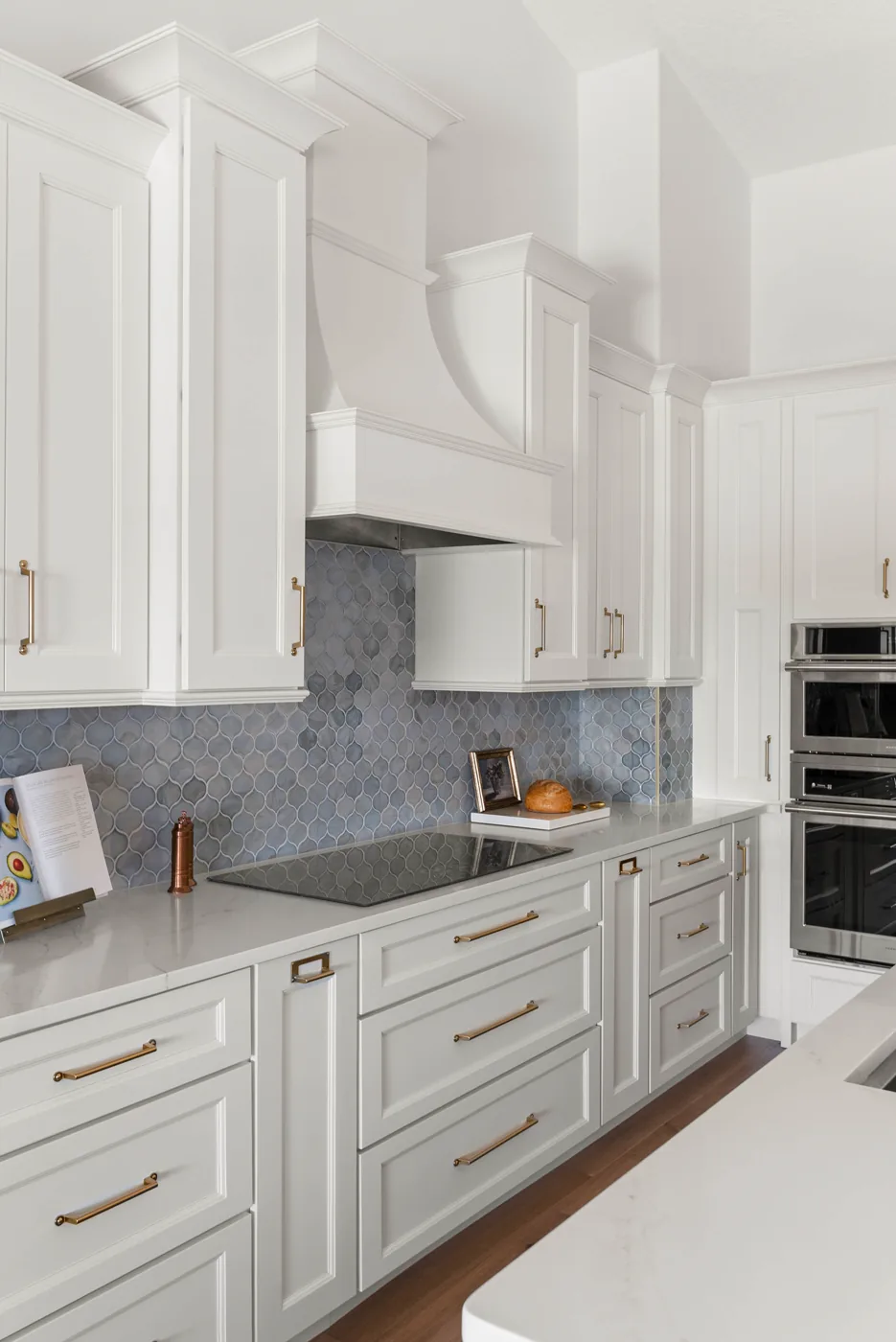 The kitchen cooking wall: a white paneled hood above a smooth-top cooktop, blue glazed arabesque tile carried to the underside of the uppers, and brass bar pulls on white recessed-panel cabinetry