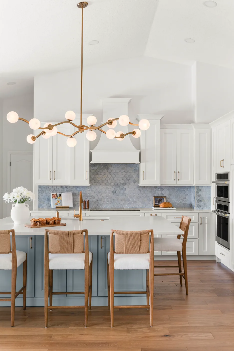 The Clermont kitchen island in soft blue, seating four under a brass orb chandelier, with the white paneled hood and blue arabesque backsplash on the cooking wall behind it