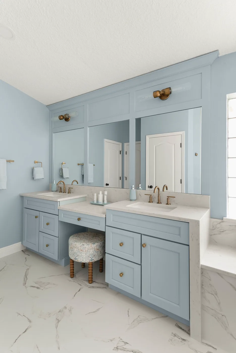The seated makeup vanity between the two blue vanities, with a floral upholstered stool, brass knobs, and a brass sconce above the framed mirror