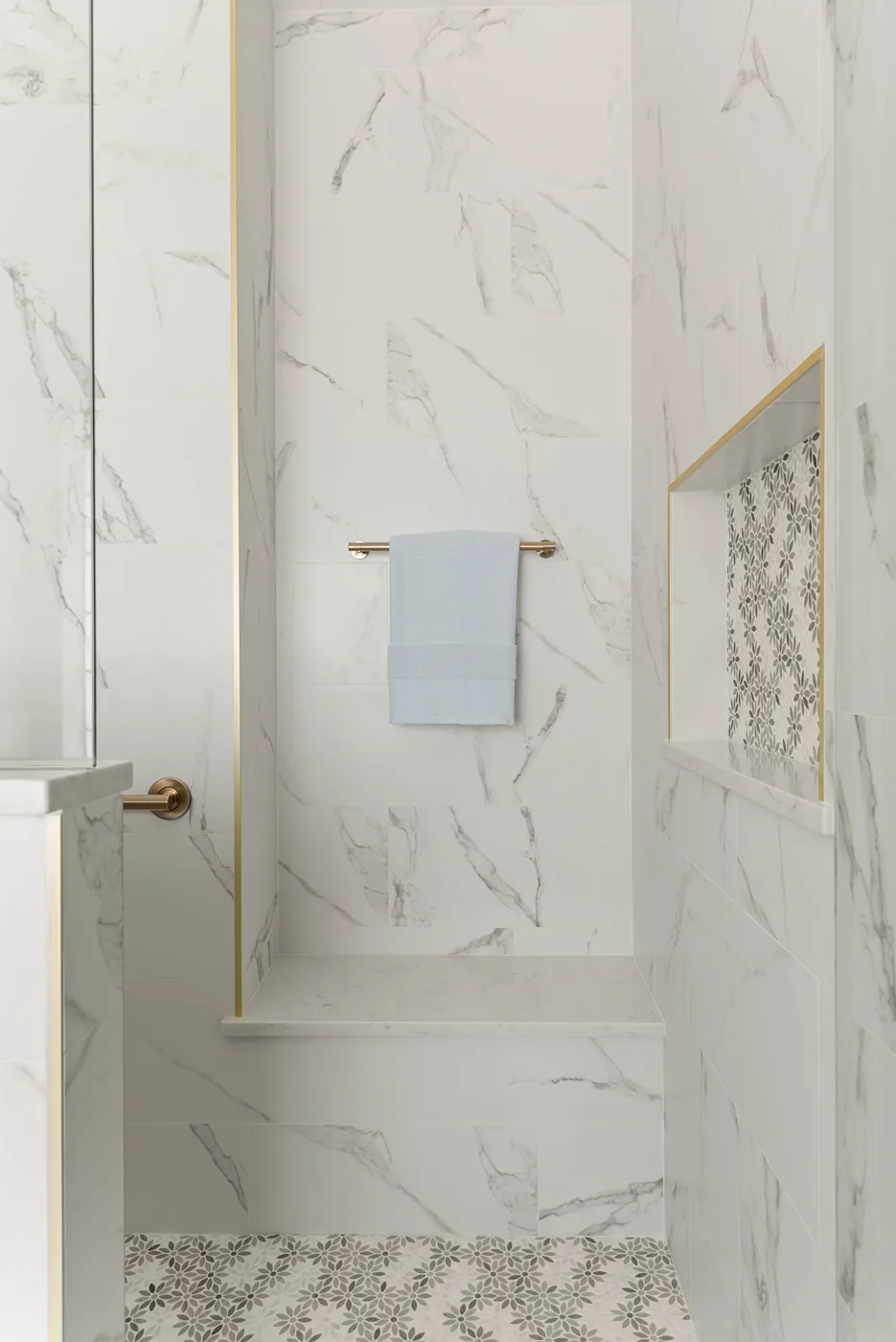 The shower built-in bench and recessed niche, the niche lined in floral mosaic and trimmed in brass, against marble-look porcelain walls