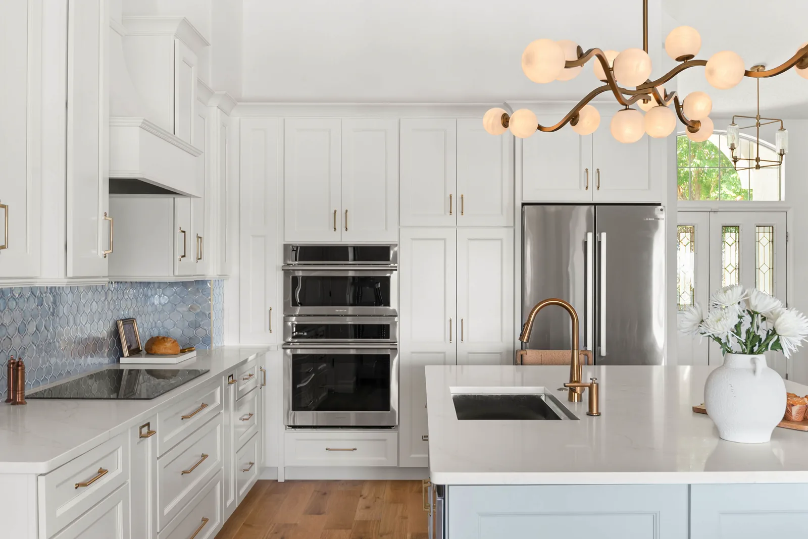 The kitchen appliance wall: a stacked stainless wall oven and microwave set into white floor-to-ceiling cabinetry beside a stainless refrigerator, with the island brass faucet and undermount sink in the foreground