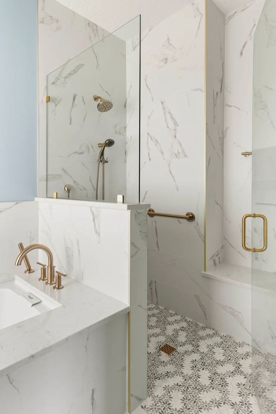 The zero-entry primary shower in large-format marble-look porcelain with a brass-trimmed edge, brass shower fittings, a grab bar, a built-in bench, and a gray-and-white floral mosaic floor