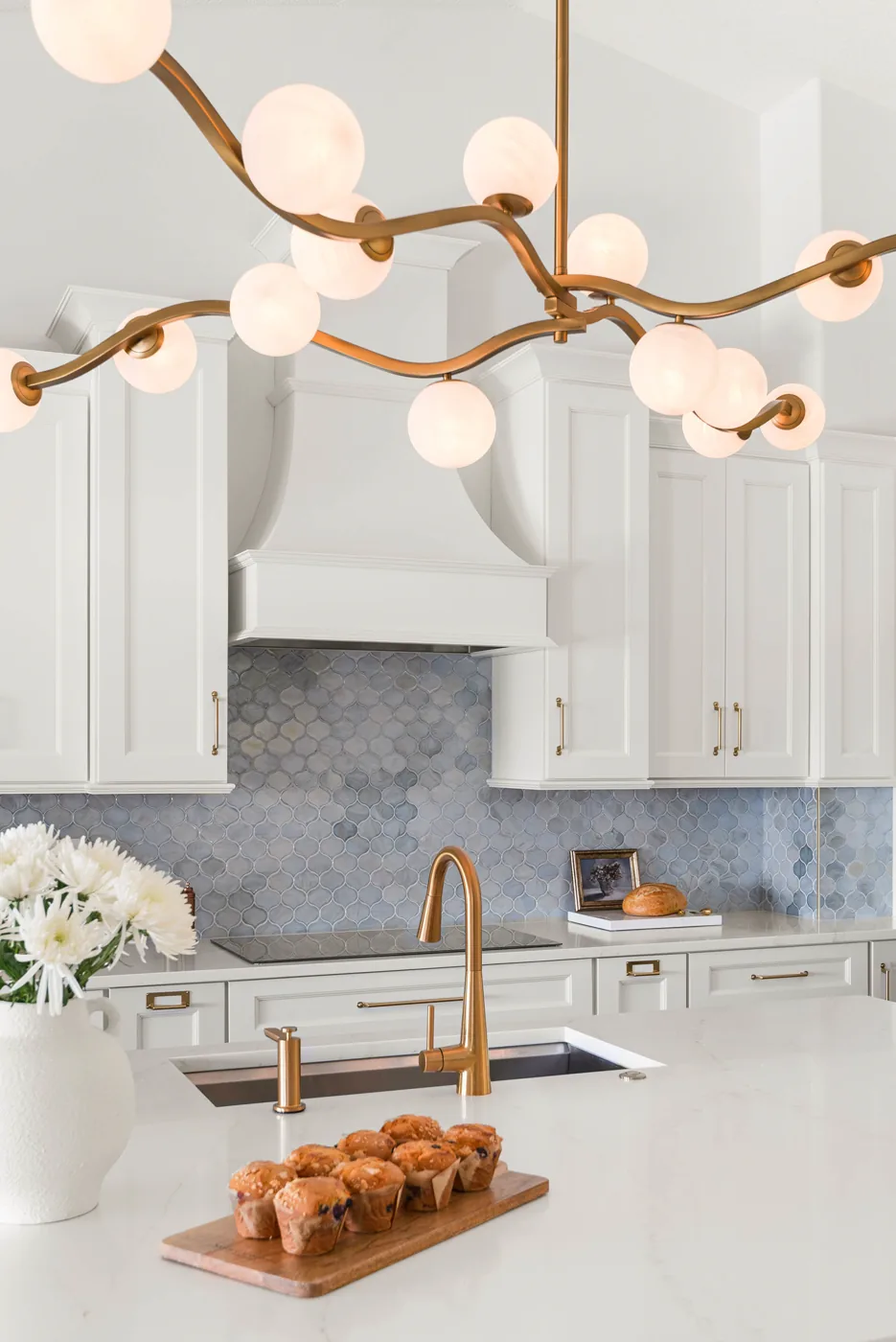 The island undermount sink and brass pull-down faucet on marble-veined counters, the brass orb chandelier overhead and the blue arabesque backsplash beyond