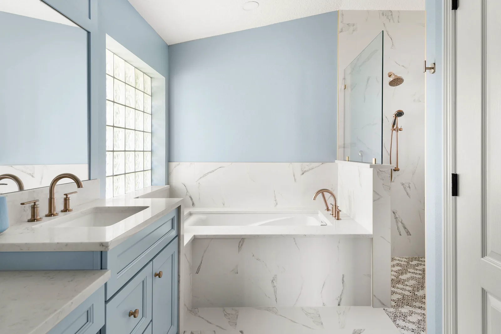 The primary bath soaking tub set into a marble-look surround below the home glass block window, with the glass shower and its patterned mosaic floor beyond