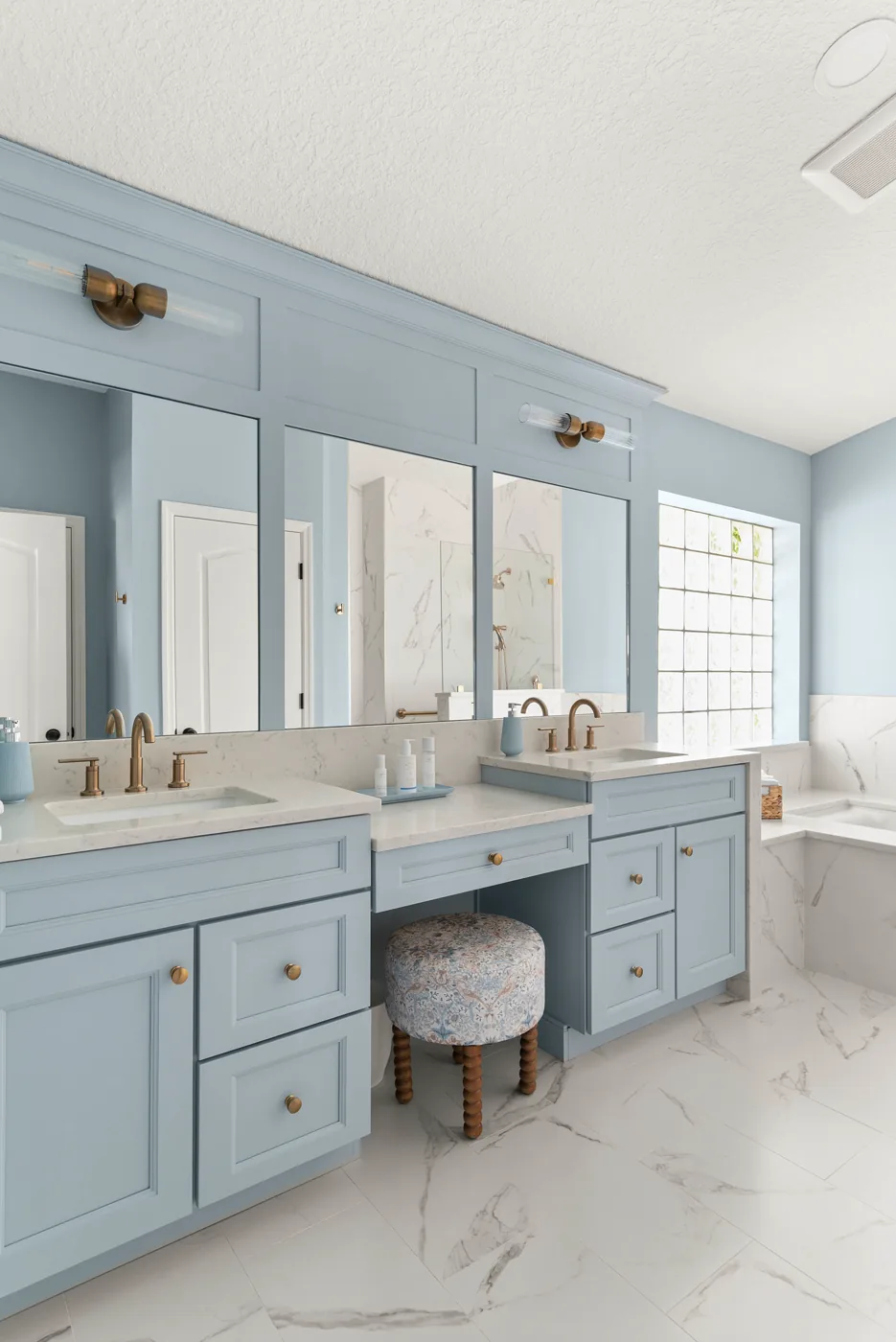The primary bath vanity wall in full: two soft blue vanities and a center makeup station with an upholstered stool, brass sconces and knobs, marble-veined counters, and the home glass block window at right