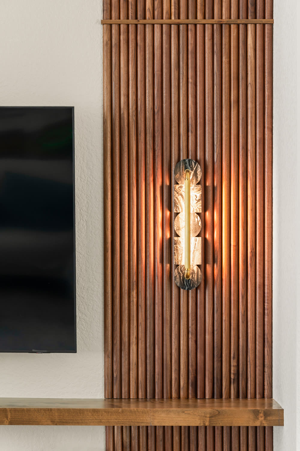 Vertical sconce with a stone shade and exposed tube bulb washing light down dark-stained wood slats beside a wall-mounted television