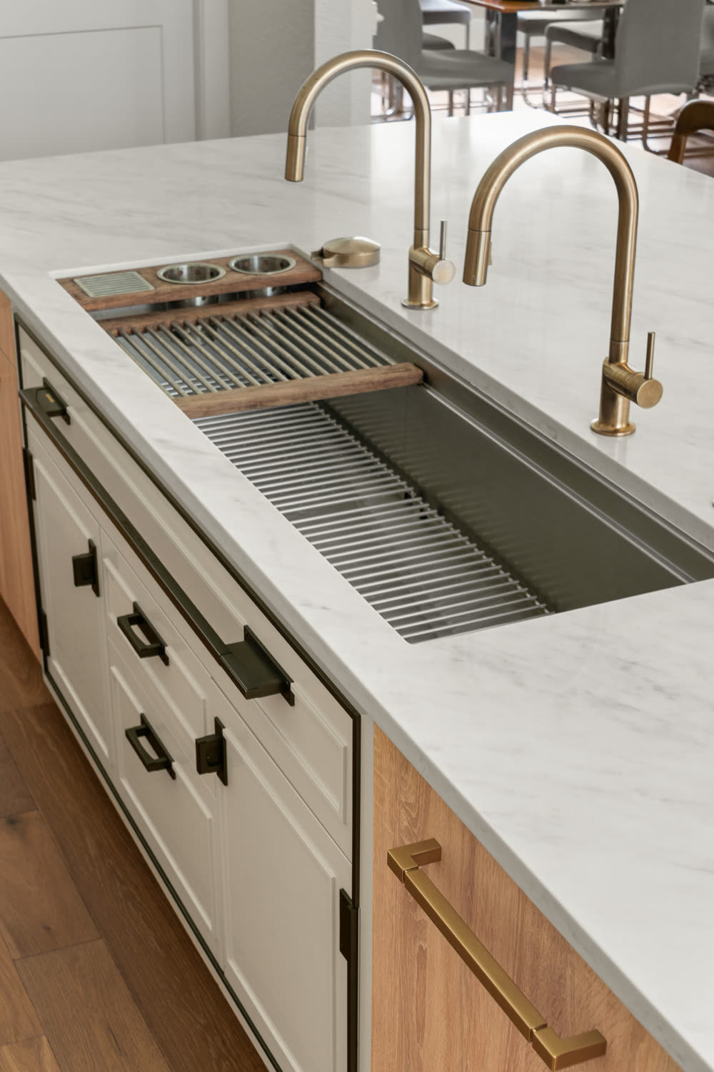 Brass faucets over a stainless workstation sink with an integrated ledge and drying rack, set into a pale veined slab island counter