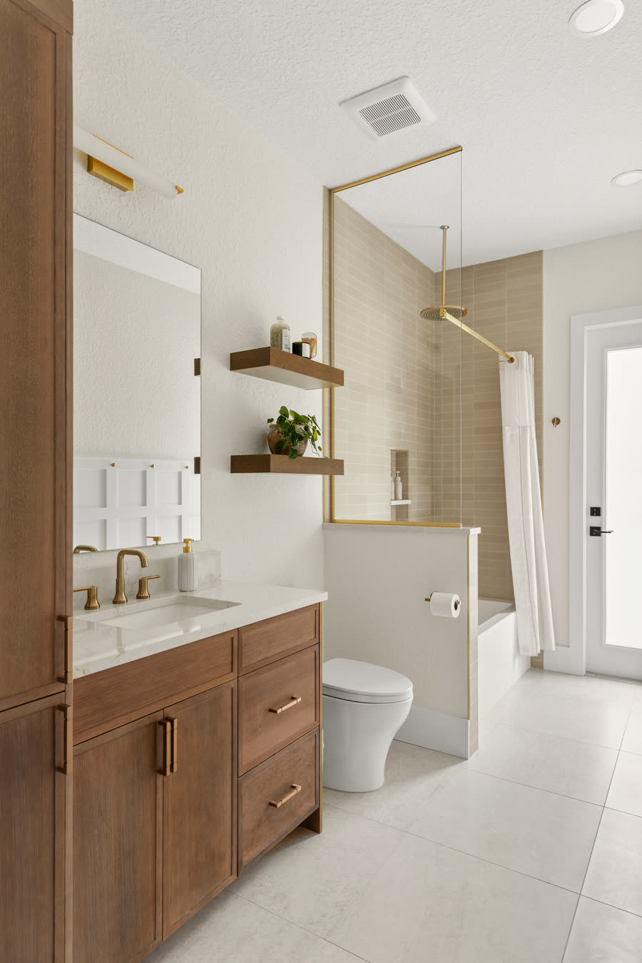 Remodeled Orlando pool bathroom with a wood vanity and full-height linen tower, brass fixtures, floating shelves and a tub-shower tiled in warm greige to the ceiling