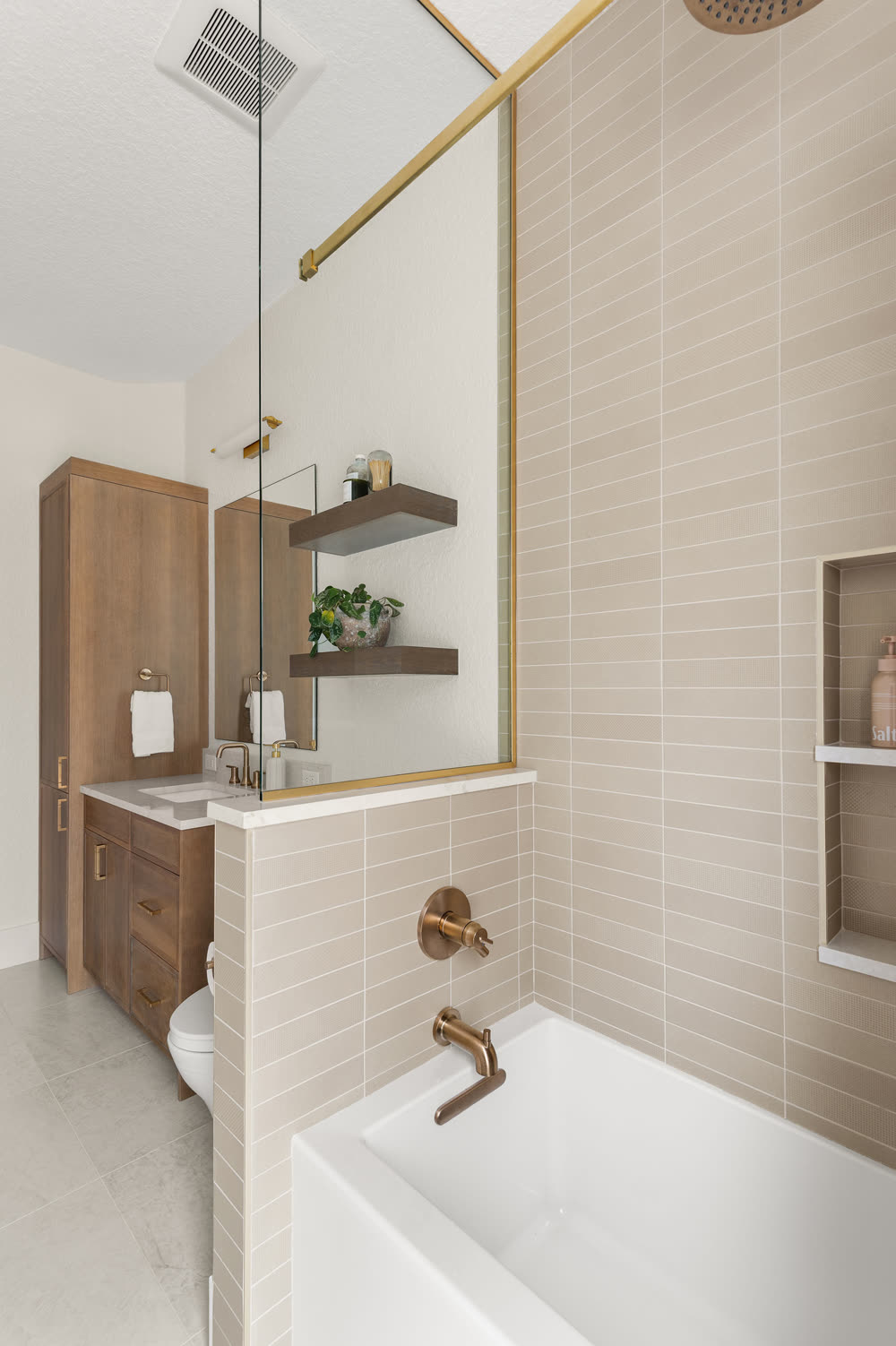 Warm greige stacked tile running to the ceiling around a white tub, with a brass tub filler, brass-framed glass panel and a recessed niche