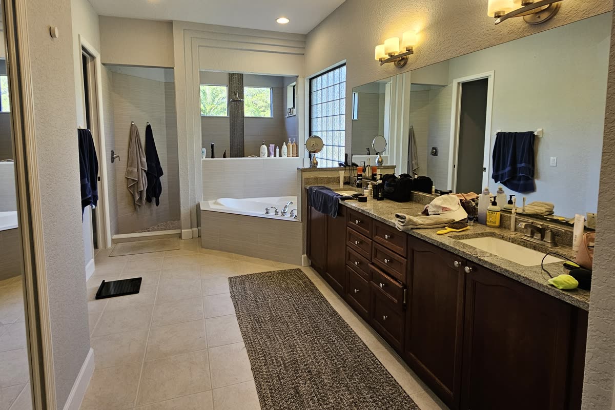 Orlando primary bathroom before remodeling, with a corner drop-in tub set on the diagonal in a tiled deck, a separate shower stall beyond it, and a dark cherry double vanity under granite and a tall glass block window