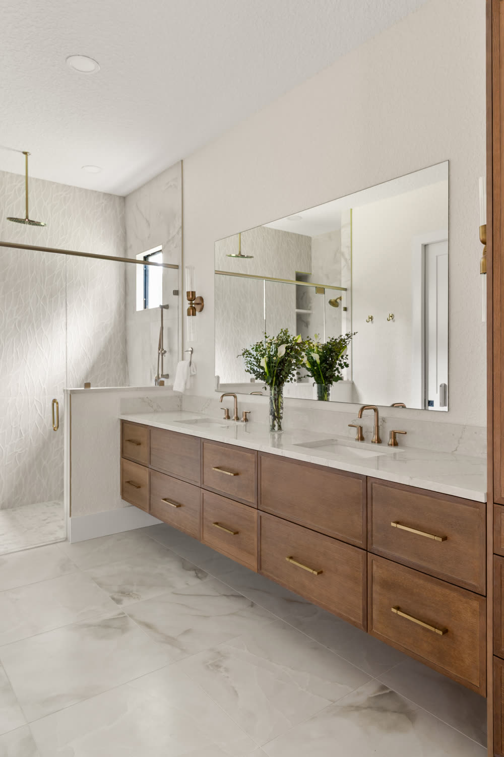 Floating wood double vanity with two undermount sinks, a full-width mirror and brass faucets in a remodeled Orlando primary bathroom, with a textured white tile shower beyond