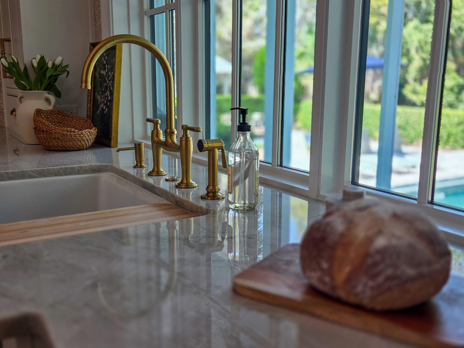 A brushed-gold bridge faucet with a side sprayer and soap dispenser at a kitchen window over a pale veined stone counter, with white tulips in a vase, a woven basket and a round loaf of bread on a wooden board, and a pool and garden visible through the windows