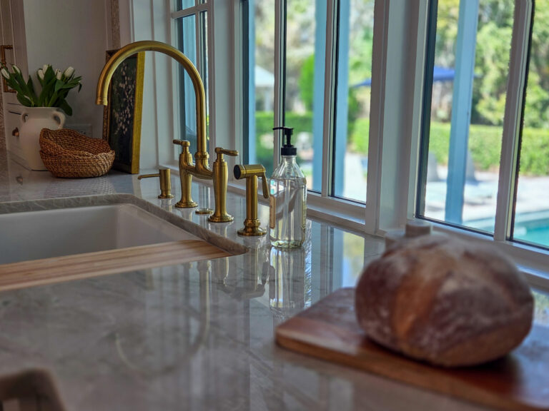 A brushed-gold bridge faucet with a side sprayer and soap dispenser at a kitchen window over a pale veined stone counter, with white tulips in a vase, a woven basket and a round loaf of bread on a wooden board, and a pool and garden visible through the windows