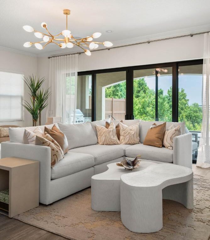 A KBF Design Gallery interior design living room with a pale sectional, sculptural white coffee tables, tan and cream pillows, a brass chandelier, and sliding glass doors opening to a covered patio