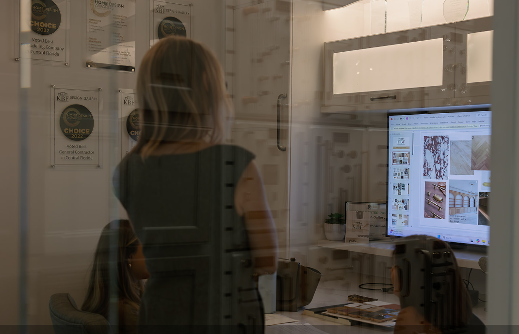 A design consultation in the KBF Design Gallery showroom in Maitland, seen through the glass wall of a design office: a KBF designer stands beside a seated homeowner at a large screen showing a selections board of veined marble, herringbone wood flooring, brass hardware and an arched vanity elevation, with tile and hardware samples on the desk and Home Design Awards plaques on the wall