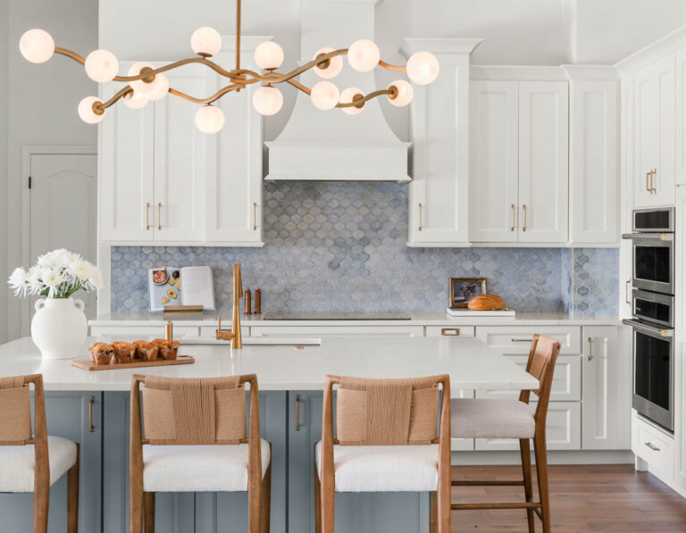 A finished KBF Design Gallery kitchen with white shaker cabinets, a blue-gray arabesque tile backsplash, a brass sculptural globe chandelier, and a blue island with woven-back wood counter stools