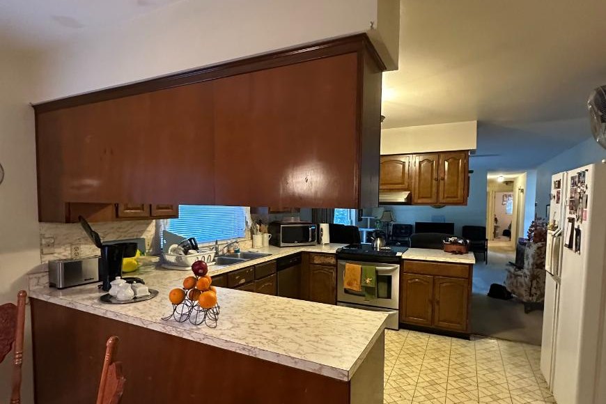 The original kitchen before the remodel, seen from the family room: a raised laminate peninsula with dark wood cabinets hung from the soffit above it, and a pass-through to the sink window beyond